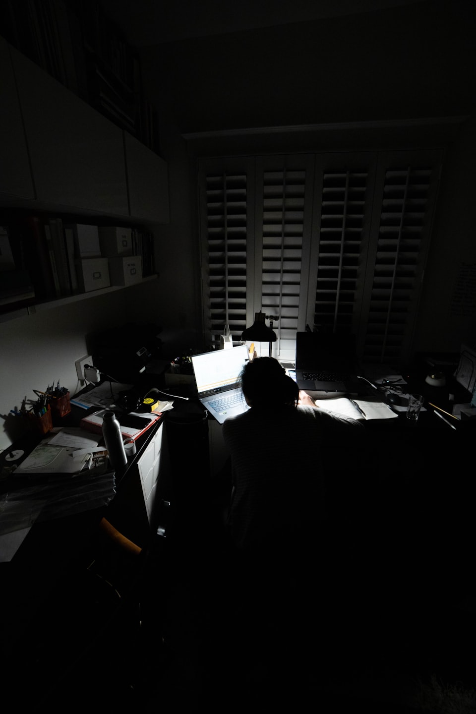 Person working on laptop at a cluttered desk in a dark room.